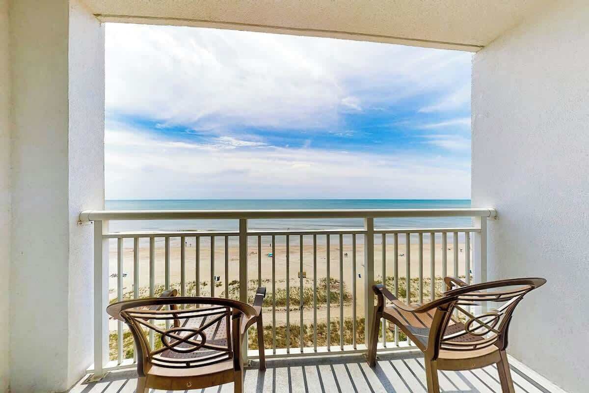A balcony with two chairs and a table, overlooking a beach with a clear sky.