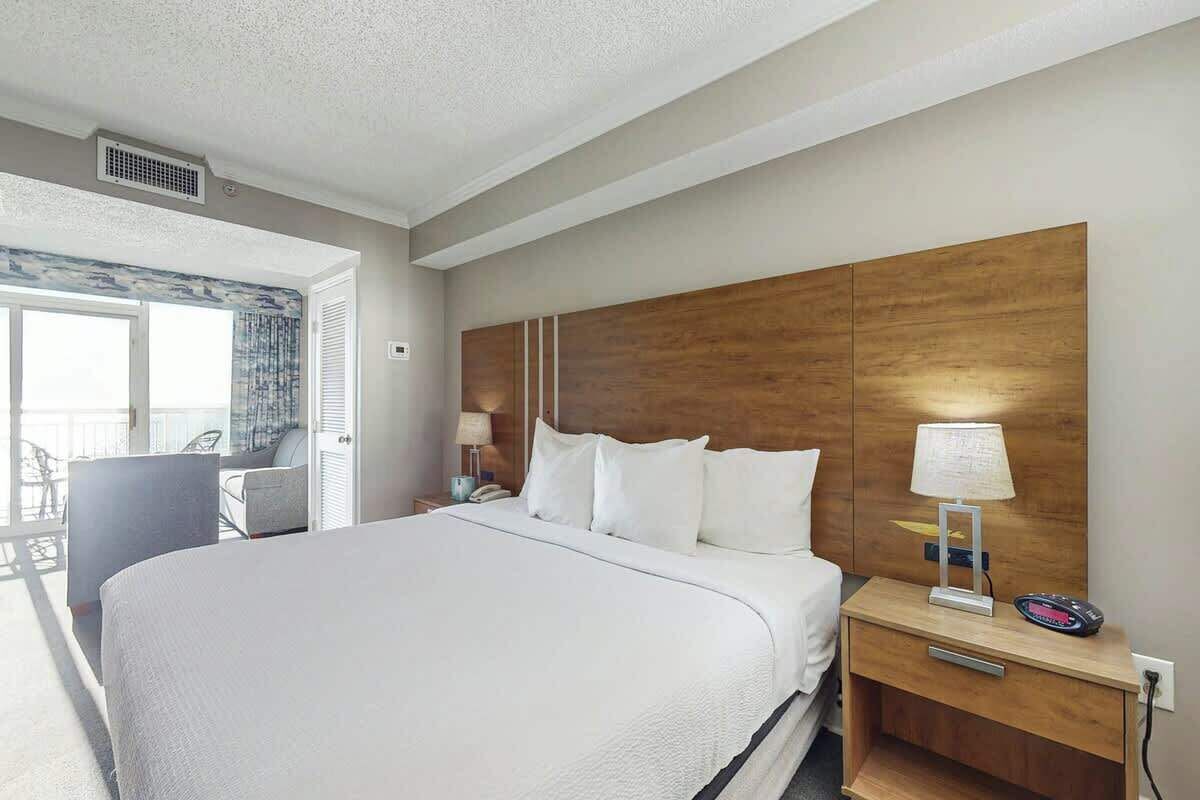 A hotel room with a large bed, a wooden headboard, a bedside table with a lamp, and a view of the balcony.
