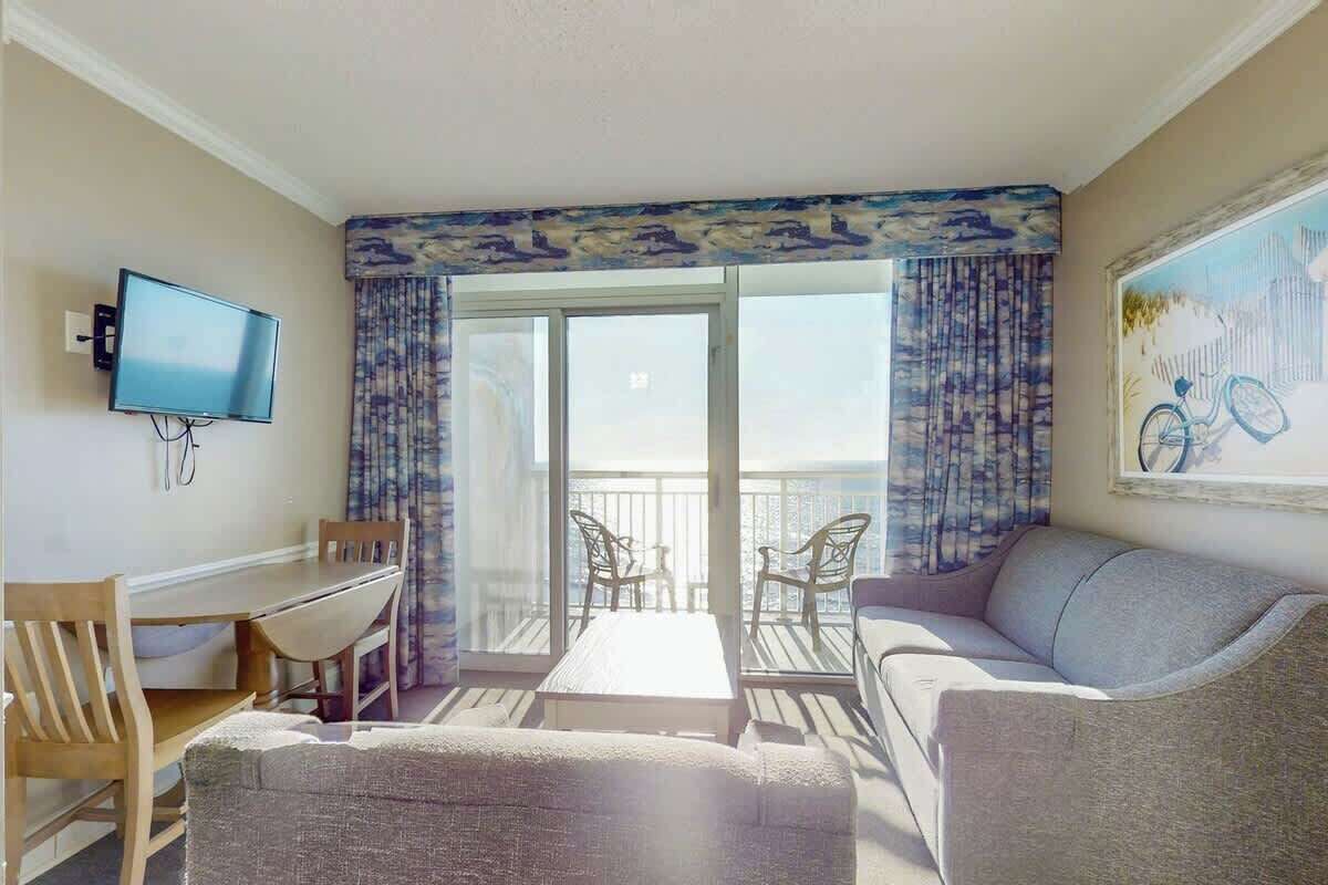 Quarto com televisão, mesa de jantar, cadeiras, sofá e varanda com vista.