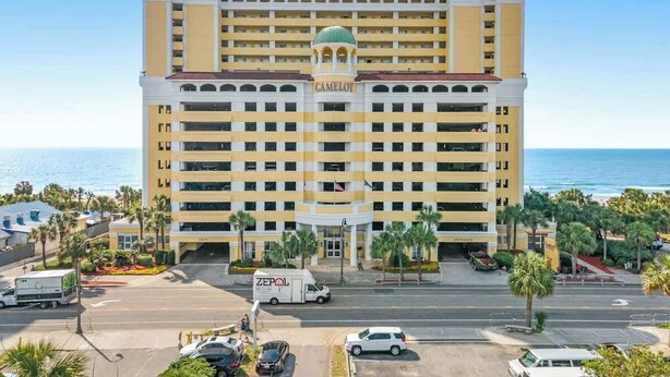 A multi-story hotel with a central entrance, surrounded by palm trees and a view of the ocean. - Camelot by the Sea 0516 (Myrtle Beach)