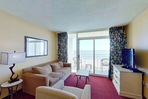 Ein Hotelzimmer mit einer Couch, einem Fernseher, einem kleinen Tisch, einer Lampe und Meerblick durch eine Schiebetür.