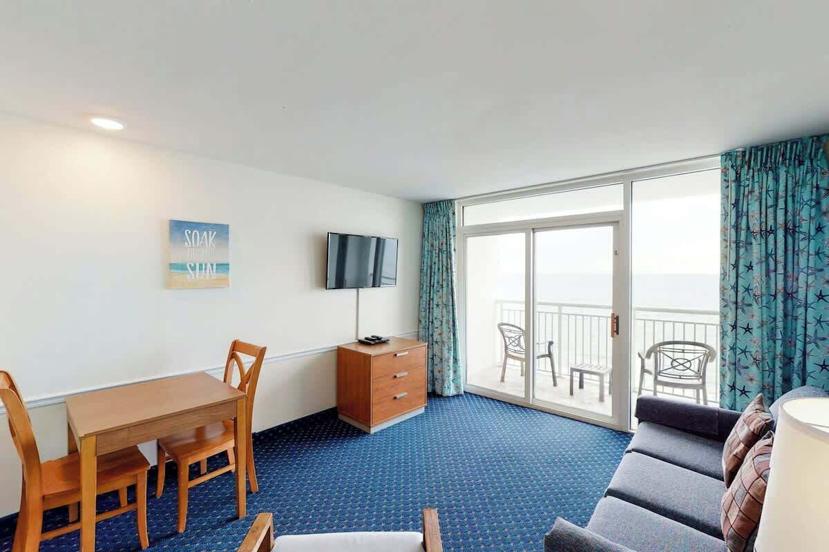 Ein Hotelzimmer mit Esstisch, Stühlen, Fernseher, Sofa und einem Balkon mit Vorhängen.