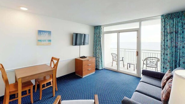 A hotel room with a dining table, chairs, a TV, a sofa, and a balcony with curtains. - Camelot by the Sea 1102 (Myrtle Beach)