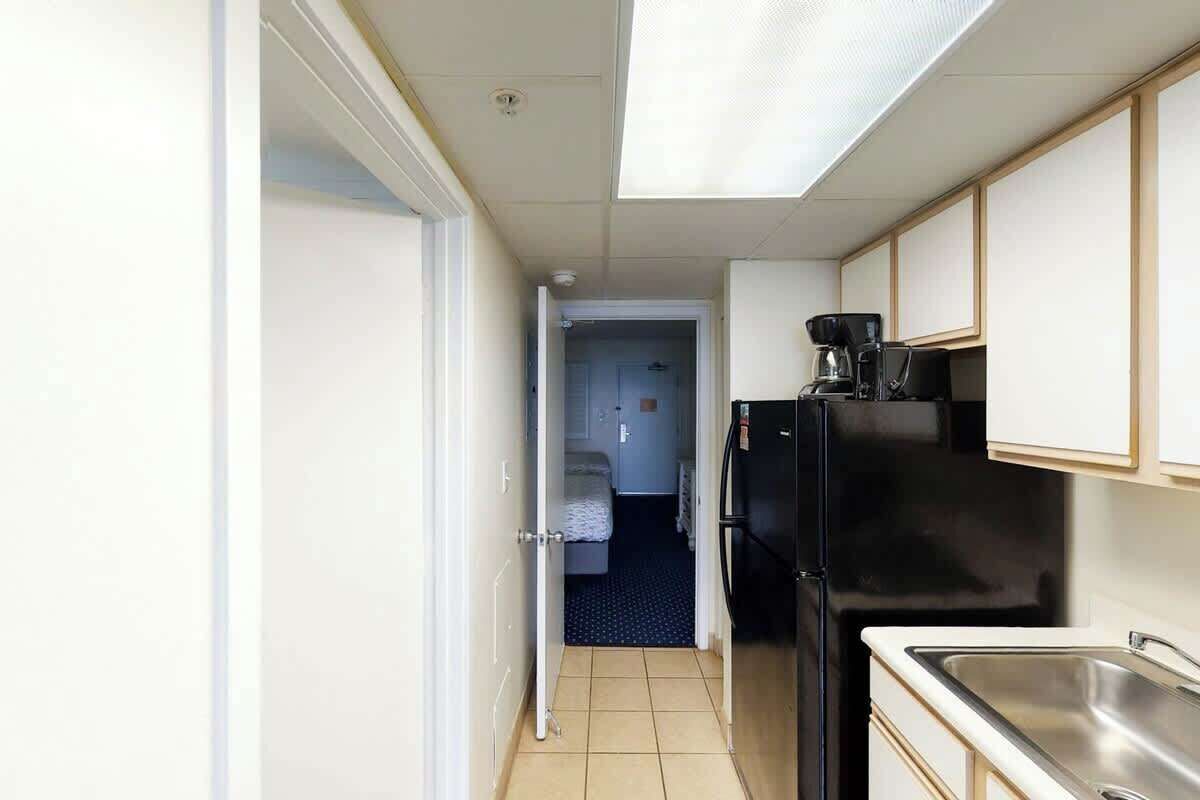 A narrow hallway leading to a bedroom with a bed and a door. A black refrigerator and a coffee maker are visible in the kitchen area.