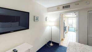 A hotel room with a bed, a TV, a desk, a lamp, and a view of the hallway. - Camelot by the Sea 1102 (Myrtle Beach)