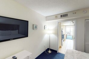 A hotel room with a bed, a TV, a desk, a lamp, and a view of the hallway. - Camelot by the Sea 1102 (Myrtle Beach)