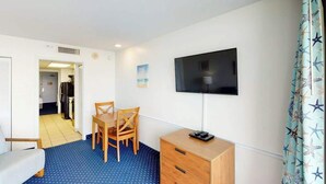 A room with a bed, a TV mounted on the wall, a wooden dresser, and a dining table with chairs. - Camelot by the Sea 1102 (Myrtle Beach)