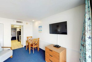 A room with a bed, a TV mounted on the wall, a wooden dresser, and a dining table with chairs. - Camelot by the Sea 1102 (Myrtle Beach)