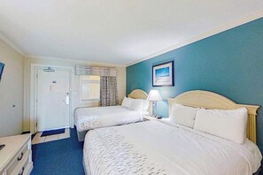 Quarto de hotel com duas camas, uma mesinha de cabeceira, um abajur, uma porta e uma janela com cortinas.