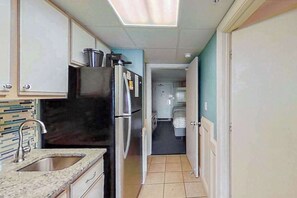 Eine kleine Küche mit Spüle, Kühlschrank und Schränken. Ein Flur führt zu einem Schlafzimmer mit einem Bett.