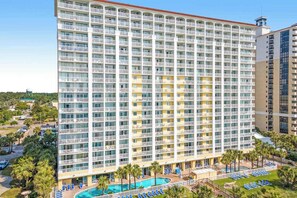 A multi-story hotel with balconies, a swimming pool, and a landscaped area with palm trees. - Camelot by the Sea 1016 (Myrtle Beach)