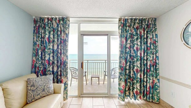 Ein Zimmer mit einer Couch, einem kleinen Tisch und Blick durch eine Schiebetür auf einen Außenbalkon mit Stühlen.