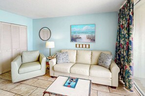 A living room with a beige sofa, a small table with a magazine, a lamp, and a painting of a beach scene. - Camelot by the Sea 1203 (Myrtle Beach)