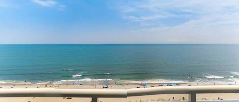 A beach view from a balcony with a railing, ocean, and beachgoers.