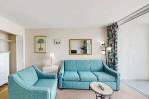 A room with a sofa, a chair, a small table with a remote, a lamp, a mirror, and a window with curtains. - Camelot by the Sea 1214 (Myrtle Beach)