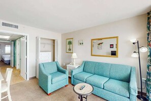 A room with a teal sofa, two matching armchairs, a small round table with a remote control, a lamp, and a mirror with a gold frame. - Camelot by the Sea 1214 (Myrtle Beach)