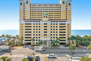 A multi-story hotel with a prominent entrance, a parking area, and a view of the ocean. - Camelot by the Sea 1214 (Myrtle Beach)