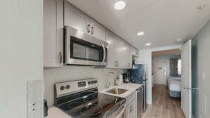 A modern kitchen with stainless steel appliances, a double sink, and a view into the adjacent bedroom. - Camelot by the Sea 1207 (Myrtle Beach)