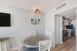 A dining area with a round table and chairs, a painting on the wall, and a kitchen in the background. - Camelot by the Sea 1207 (Myrtle Beach)