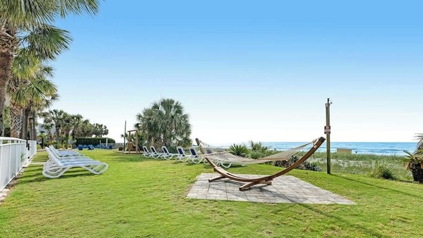 A grassy area with a hammock and lounge chairs, a white fence, and a view of the ocean. - Camelot by the Sea 1207 (Myrtle Beach)