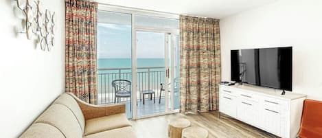 A room with a sofa, a television, a wooden cabinet, and a view of the sea through a glass door.