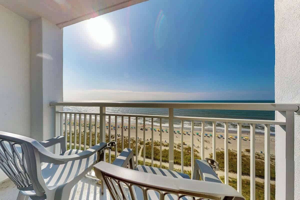 A balcony with a view of a beach, a table, and two chairs.