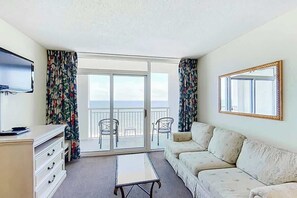 Ein Zimmer mit einer Couch, einem Fernseher, einem Tisch und einem Balkon mit Stühlen.