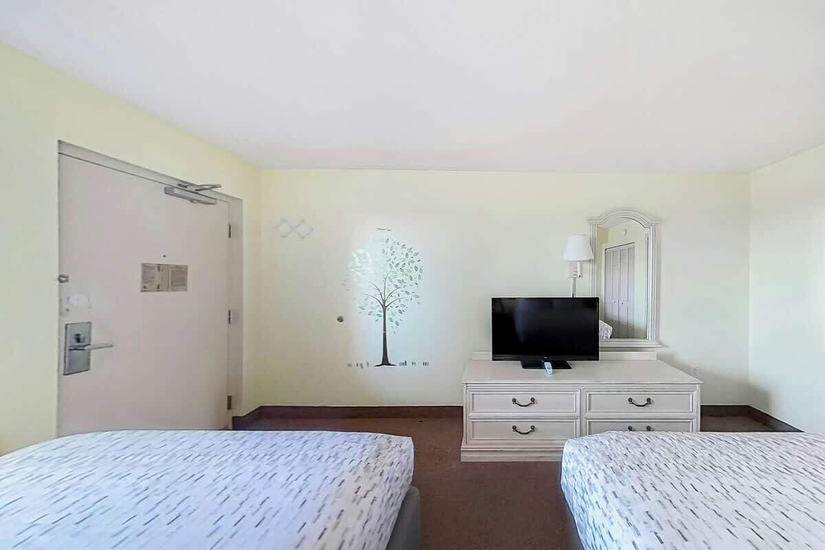 Une chambre d’hôtel avec deux lits, un téléviseur posé sur une commode, une porte avec une poignée et une décoration murale représentant un arbre.