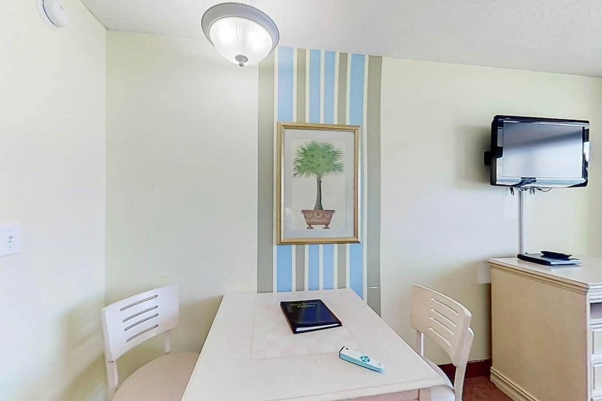 A room with a white table, two white chairs, a framed picture of a tree, a television on a stand, and a small cabinet.