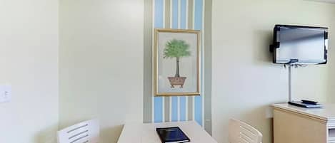 A room with a white table, two white chairs, a framed picture of a tree, a television on a stand, and a small cabinet.