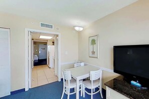 A room with a dining table, chairs, a flat-screen TV, and a kitchen area visible through an open door. - Camelot by the Sea 1512 (Myrtle Beach)