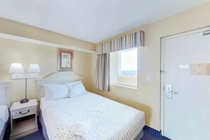 A hotel room with a bed, bedside lamps, a window with curtains, and a door. - Camelot by the Sea 1512 (Myrtle Beach)