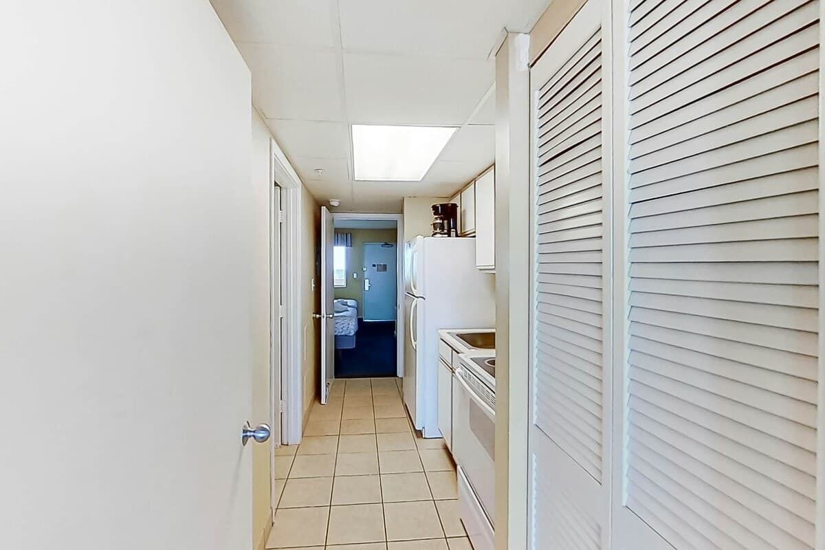 A narrow hallway with a tiled floor, a door on the left, and a kitchen area with appliances on the right.