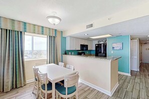 Une cuisine compacte dotée d’un îlot central, d’un coin repas avec quatre chaises et d’une vue sur l’extérieur à travers une fenêtre agrémentée de rideaux.