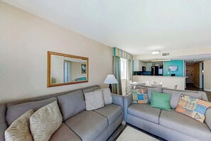 A living room with two sofas, a mirror, and a kitchen area in the background. - Camelot by the Sea 1601 (Myrtle Beach)