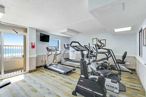 A gym with treadmills, elliptical machines, and a stationary bike, with a view of the sea through a sliding door. - Camelot by the Sea 1514 (Myrtle Beach)