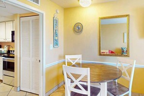A dining area with a round table, white chairs, a wall clock, a mirror, and a kitchen in the background. - Camelot by the Sea 1804 (Myrtle Beach)