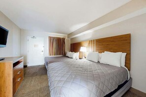 A hotel room with a large bed, a wooden headboard, a desk with drawers, a television, and a door with striped curtains. - Camelot by the Sea 1607 (Myrtle Beach)