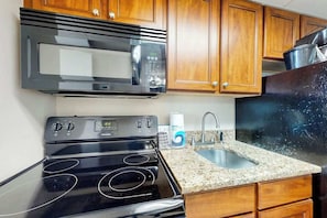 A kitchen with a microwave, stove, granite countertop, wooden cabinets, and a sink. - Camelot by the Sea 1607 (Myrtle Beach)