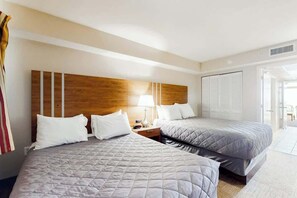 A hotel room with two beds, a wooden headboard, a nightstand, and a window with blinds. - Camelot by the Sea 1607 (Myrtle Beach)