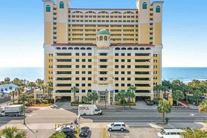 A multi-story hotel with a central entrance, surrounded by palm trees and a view of the ocean. - Camelot by the Sea 1607 (Myrtle Beach)