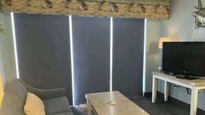 A living room with a sofa, a coffee table, a television on a stand, and a large window with blinds. - Camelot by the Sea 1603 (Myrtle Beach)