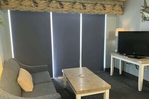 A living room with a sofa, a coffee table, a television on a stand, and a large window with blinds. - Camelot by the Sea 1603 (Myrtle Beach)