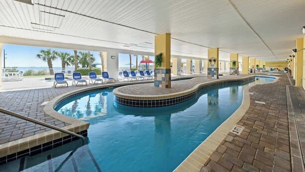 A swimming pool with a curved design, surrounded by lounge chairs and a view of the beach. - Camelot by the Sea 1603 (Myrtle Beach)
