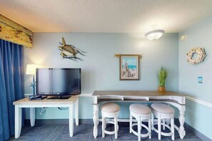 A room with a TV on a white table, a framed picture on the wall, and a plant on a wooden bench. - Camelot by the Sea 1603 (Myrtle Beach)