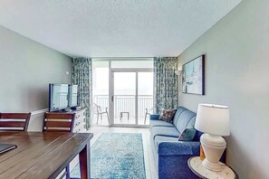 A room with a dining table, a sofa, a TV, a lamp, and a view of the outdoors through a sliding door. - Camelot by the Sea 1809 (Myrtle Beach)