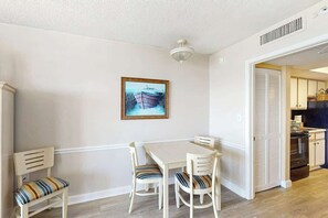 Un coin repas avec une table et des chaises, un tableau accroché au mur et une cuisine visible à travers une porte ouverte.