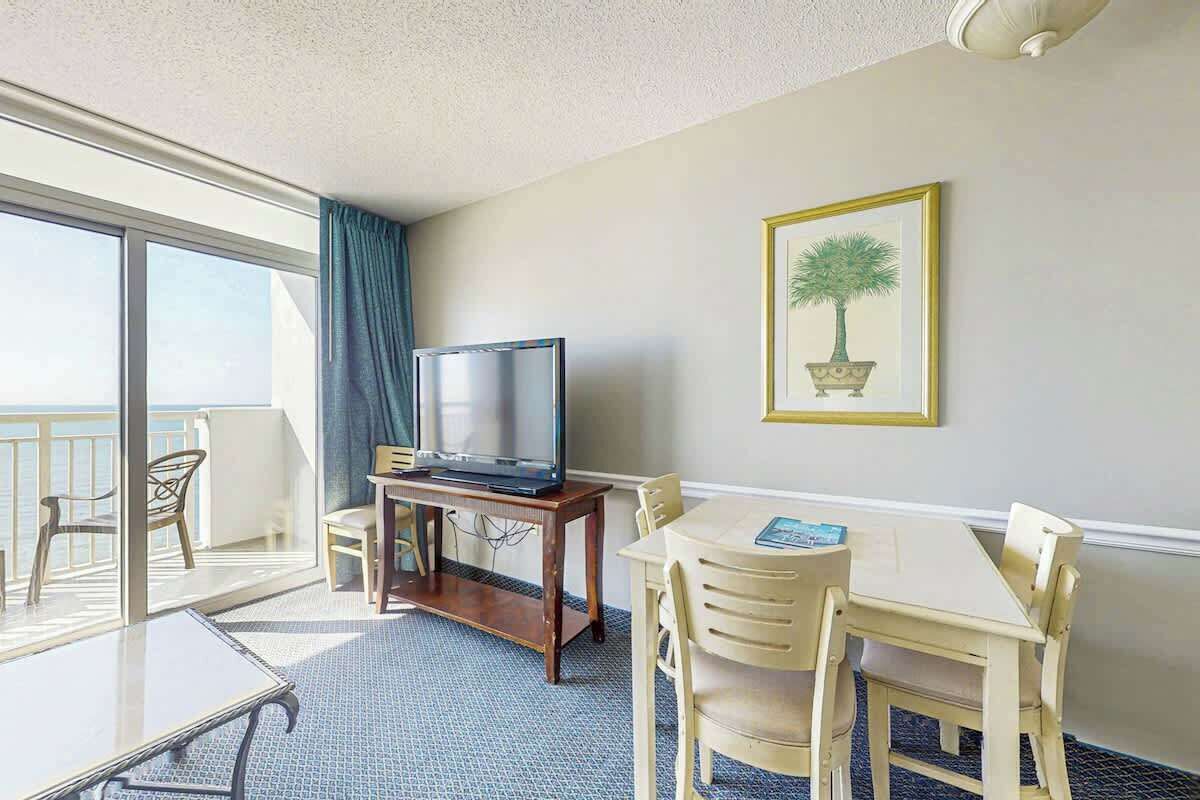 A hotel room with a balcony, a television on a wooden stand, a dining table with chairs, and a framed picture on the wall.