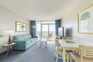 A hotel room with a sofa, a small table, a TV, and a balcony with chairs. - Camelot by the Sea 1711 (Myrtle Beach)