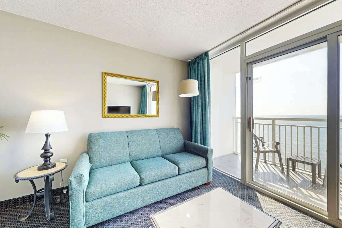 A hotel room with a sofa, a table, a lamp, a mirror, and a balcony with a view.